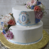 The WD-009 is a two-tiered white cake adorned with pastel flowers, a floral monogram "DM," light blue ribbons at each base, and sits on a gold board.
