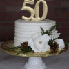 The CH-000F is a white frosted cake on a white stand, topped with a gold 50. It features white flowers, pine cones, greenery, and sits on a gold foil base against a brick wall background.