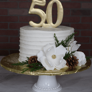 The CH-000F is a white frosted cake on a white stand, topped with a gold 50. It features white flowers, pine cones, greenery, and sits on a gold foil base against a brick wall background.