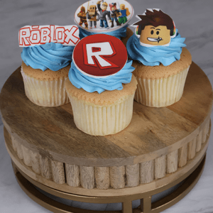 Three CC-031B Gold cupcakes with chocolate filling and blue frosting sit on a wooden stand. Each cupcake features a Roblox-themed decoration: the Roblox logo, a game character face, and a group of Roblox avatars.