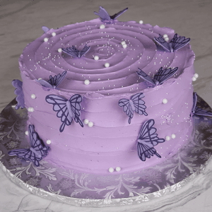 The BD-060I features a round cake with smooth light purple frosting, adorned with purple butterfly toppers, white sugar pearls, and a swirl pattern on top, presented on a silver floral-patterned cake board.