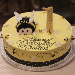 The CB-012J is a round yellow birthday cake with small bees, white flowers, a large bee face and wings, plus a number “1” topper. “Happy Birthday Kristin” is written in cursive icing.