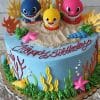 The SC-073 Baby Shark cake features blue frosting, colorful seaweed, starfish, shells, three Baby Shark figures (blue, yellow, pink), and “Happy Birthday” in red icing.