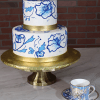 The WD-001E two-tier cake with blue floral designs and gold bands sits on a gold stand beside a matching floral teacup and saucer, all arranged on marble with a brick wall in the background.