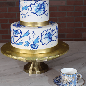 The WD-001E two-tier cake with blue floral designs and gold bands sits on a gold stand beside a matching floral teacup and saucer, all arranged on marble with a brick wall in the background.