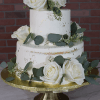 The WD-001F is a two-tiered white cake adorned with white roses and green eucalyptus leaves, displayed on a gold cake stand against a brick wall backdrop.