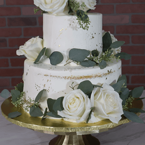 The WD-001F is a two-tiered white cake adorned with white roses and green eucalyptus leaves, displayed on a gold cake stand against a brick wall backdrop.