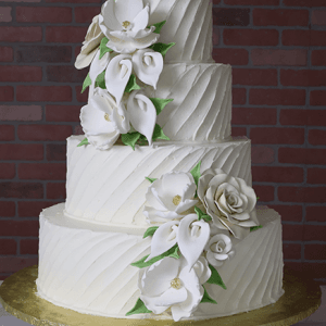 The WD-001C is a three-tiered white cake with textured diagonal frosting, adorned with large white sugar flowers and green leaves, displayed on a gold cake board against a brick wall backdrop.