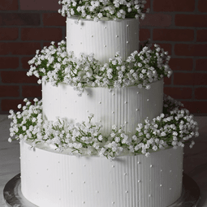 Product WD-001G: Elegant three-tiered white wedding cake adorned with small white flowers and piped dots, presented on a silver cake board against a brick wall background.