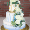 The WD-001I is a three-tiered white cake with blue and gold accents, decorated with white roses, greenery, gold ribbon borders, and set on a gold cake board against a brick wall background.