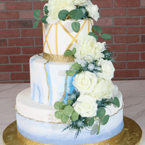 The WD-001I is a three-tiered white cake with blue and gold accents, decorated with white roses, greenery, gold ribbon borders, and set on a gold cake board against a brick wall background.