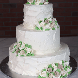 The WC-000K is a three-tiered white cake adorned with clusters of white and light brown roses and green leaves, displayed on a silver board against a brick wall.