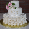 The WD-001J features two white tiers, ruffled bottom icing, a white ribbon, piped ESM initials, pink and white sugar roses on top, all set on a gold board with a brick backdrop.