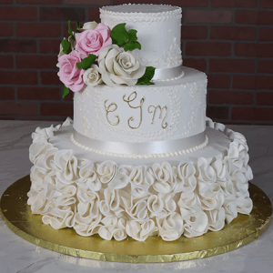 The WD-001J features two white tiers, ruffled bottom icing, a white ribbon, piped ESM initials, pink and white sugar roses on top, all set on a gold board with a brick backdrop.
