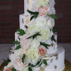 The WD-001L is a four-tier white cake with gold leaf accents, adorned with cascading white and blush pink roses, peonies, and green leaves, set against a brick wall background.