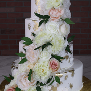The WD-001L is a four-tier white cake with gold leaf accents, adorned with cascading white and blush pink roses, peonies, and green leaves, set against a brick wall background.