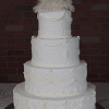 The WD-001M is a four-tiered white wedding cake with ornate frosting designs and a bride-and-groom figurine topper, displayed against a brick wall background.