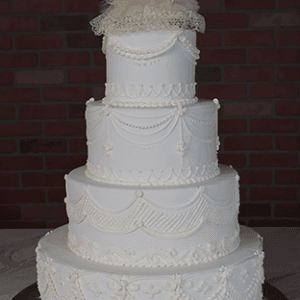 The WD-001M is a four-tiered white wedding cake with ornate frosting designs and a bride-and-groom figurine topper, displayed against a brick wall background.
