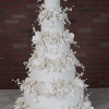 The WD-001N is a tall, elegant five-tier white wedding cake adorned with intricate sugar flowers and leaves, displayed on marble with a brick wall backdrop.