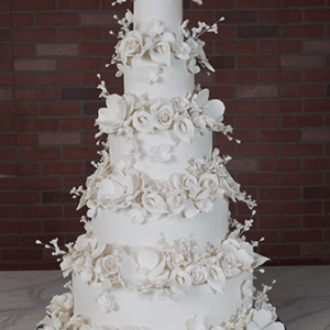 The WD-001N is a tall, elegant five-tier white wedding cake adorned with intricate sugar flowers and leaves, displayed on marble with a brick wall backdrop.