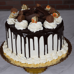The BD-018S Chocolate Peanut Butter cake features white frosting, a chocolate drip, whipped cream swirls, mini peanut butter cups, and chocolate curls on a gold board. Displayed on marble with a brick wall backdrop.