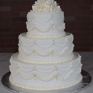 The WC-000C is a three-tiered white wedding cake with elegant piped frosting swags and white icing roses, presented on a silver cake board against a dark brick background.