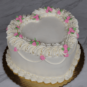 CB-036D is a heart-shaped white frosted cake with piped white icing and small pink and green flower accents, displayed on a gold cake board against a marble background.
