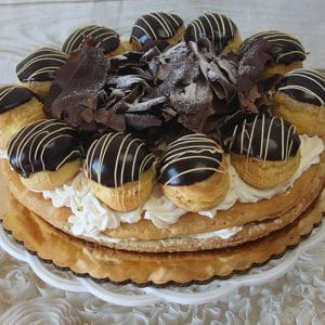 The GD-029A Gateau St. Honoré with Ganache is a round cream puff cake topped with chocolate profiteroles, chocolate shavings, and piped whipped cream, served on a gold cake board.