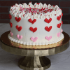 The VC-000C Valentine Cake, a round white cake with red and pink heart-shaped frosting, white swirls, and red sprinkles, is presented on a gold cake stand in front of a brick wall background.