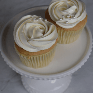 Two CC-011 chocolate cupcakes with white chocolate mousse filling and white rose-patterned frosting with silver pearls sit on a white cake stand with a beaded edge, set against a light marble-like background.