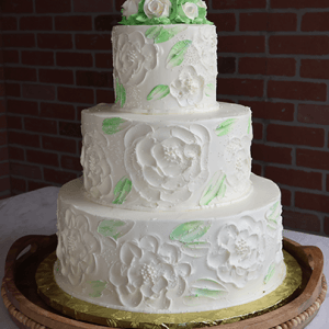 The WC-000A is a three-tiered white cake with floral designs and green leaves, topped with white roses, and displayed on a wooden tray against a brick wall background.