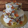 The TC-009 is a two-tiered white cake adorned with orange, yellow, and brown autumn leaves and vines, set on a gold board with a rustic wood background.