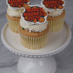 Three CC-098B March Madness Gold Cupcakes with chocolate filling and basketball-themed 2025 toppers sit on a white cake stand. The cupcakes have white frosting, and the background is a light grey marble surface.