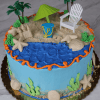 The SC-004 Beach Chair cake is round with blue icing water, crushed cookie sand, plastic palm trees, a chair, umbrella, seashells, starfish on top; green icing waves and sea plants decorate the sides.