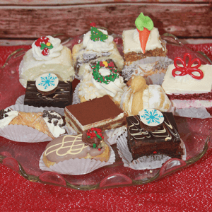 The CH-000 Assorted Mini Pastries Chef's Choice with Holiday decor features decorated mini pastries and cakes topped with festive icing and sprinkles, beautifully arranged on a glass plate on a red textured surface.