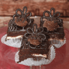 Three Hazelnut Truffle Squares, filled with cream and topped with chocolate frosting, curls, and flower decorations, are displayed on a glass plate against a rustic wooden backdrop. One square has a bite taken out.