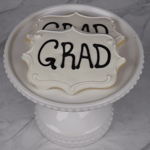 Two white frosted cookies with "GRAD" in black icing (Product: GC-0004) are displayed on a white decorative cake stand against a marble background.