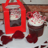 The VC-042 Individual Red Velvet cupcake, topped with white frosting, red heart sprinkles, and a red Love topper, sits on a white tray with rose petals, next to a matching cupcake in a red windowed box.