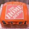 CO-009: A square orange cake with The Home Depot logo centered, icing tools on the sides, and "Hi! My name is Phillip and I'm 80." written on top, displayed on a silver surface.