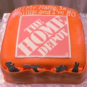 CO-009: A square orange cake with The Home Depot logo centered, icing tools on the sides, and "Hi! My name is Phillip and I'm 80." written on top, displayed on a silver surface.