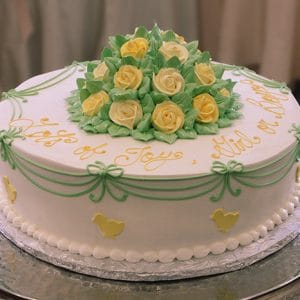 The BB-010 is a round white cake with yellow and green icing flowers on top, green bows around the sides, small yellow duck shapes, and a cursive message in yellow icing.