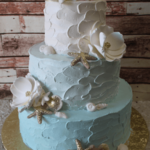 TC-015: Two-tiered cake with textured blue and white frosting, adorned with white sugar flowers, gold starfish, and white seashells on a gold cake board, set against a rustic wooden background.