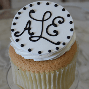 A gold cupcake with chocolate mousse filling, topped with a round white fondant disc decorated with black dots and the initials ALE written in black icing.