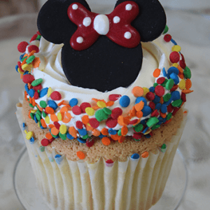 A CC-127 Gold cupcake with chocolate mousse filling, topped with white frosting, colorful round sprinkles, and a Minnie Mouse-shaped chocolate decoration with a red bow and white polka dots sits on a clear glass plate.