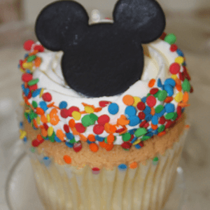 A gold cupcake, filled with chocolate mousse, is topped with white frosting, colorful confetti sprinkles, and a black decoration shaped like Mickey Mouse's head.