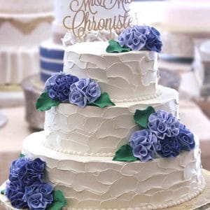 The WD-283 is a three-tiered white wedding cake with textured icing, purple flower accents, green leaves, and a gold "Mr & Mrs Christophe" topper. Other cakes appear blurred in the background.