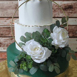 WD-008 is a three-tier cake with white roses, green succulents, eucalyptus leaves, a gold hoop, and gold trim on the bottom tier. It sits on a gold board against a wooden plank background.