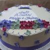 The BR-026 is a round cake with purple, red, and green flower-shaped fondant, a purple ribbon at the base, and "Best Wishes Marigo & Stefanos" written in blue icing on top.