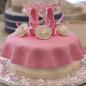 The CB-026C is a round cake covered in pink fondant with ruffled edges, topped with two pink fondant ballet slippers and three white fondant roses, all displayed on a decorative silver platter.