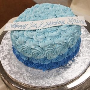 The BD-062 Blue rosette is a round cake with two shades of blue rosette frosting, topped with a white "Happy 1st Birthday Yaim" strip, and displayed on a silver-patterned cake board.